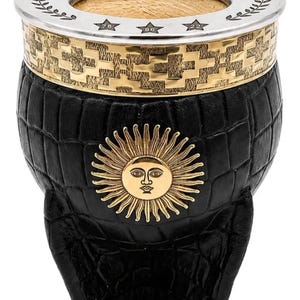 Può includere: Una zucca di yerba mate nera con un emblema solare dorato e una fascia decorativa dorata. Il bordo è argentato con le parole "CORONADOS DE GLORIA". La zucca ha un motivo strutturato a forma di coccodrillo.