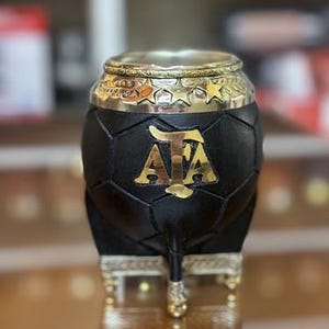 Puede incluir: Una calabaza de yerba mate negra con borde y base dorados. La calabaza tiene un diseño de balón de fútbol y un emblema "AFA" dorado. El borde está decorado con estrellas. La base tiene patas doradas ornamentadas.