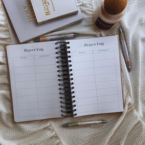 May include: An open spiral-bound "Prayer Log" notebook with lined pages, next to a closed book titled "HOLY BIBLE". A pen rests on the right side of the notebook. A candle and another pen are also visible.