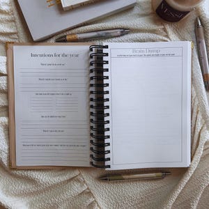 May include: Open journal with spiral binding, featuring the text "Intentions for the year" and "Brain Dump" on facing pages. The journal is open to lined pages with prompts. Pens and a candle are visible in the background.