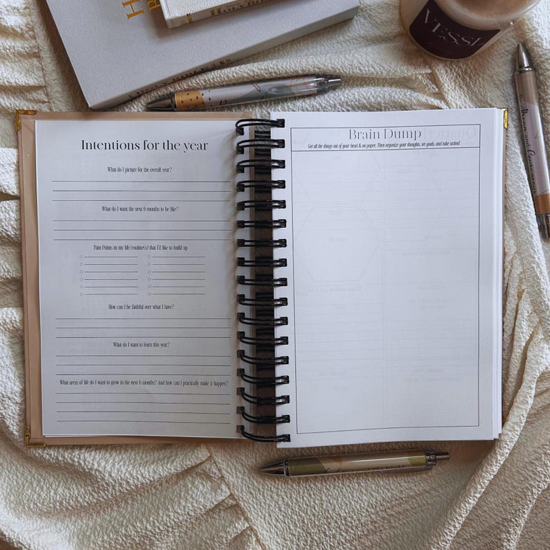 May include: Open journal with spiral binding, featuring the text "Intentions for the year" and "Brain Dump" on facing pages. The journal is open to lined pages with prompts. Pens and a candle are visible in the background.