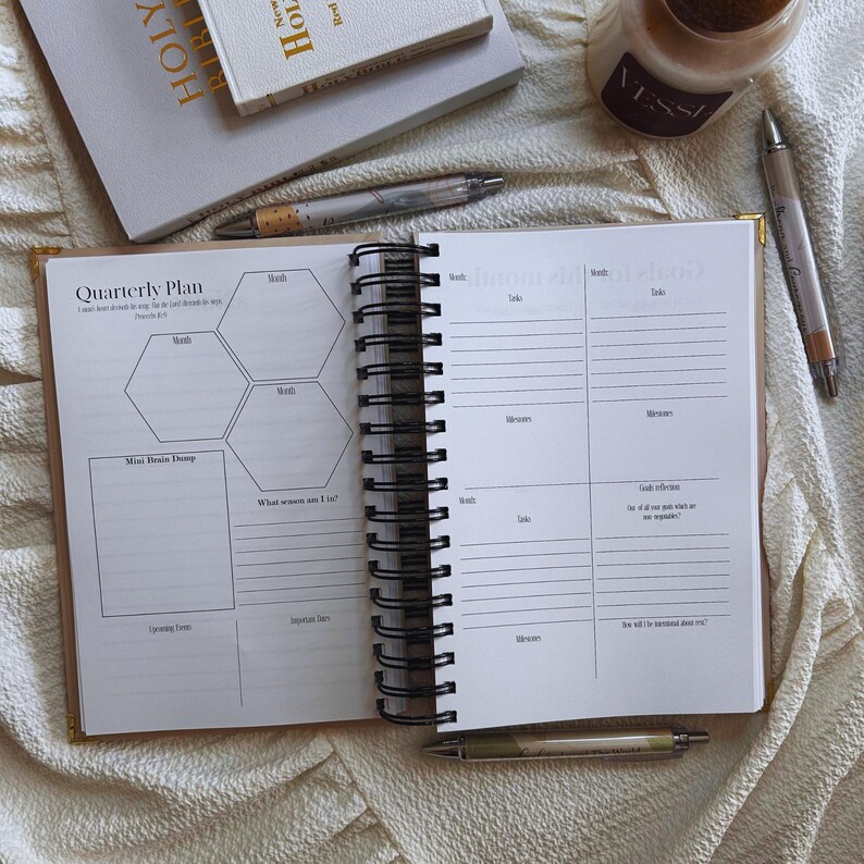 May include: Open spiral-bound planner with a quarterly plan layout, featuring sections for months, tasks, and notes. A pen rests on the open pages. A small stack of books and a candle are in the background.