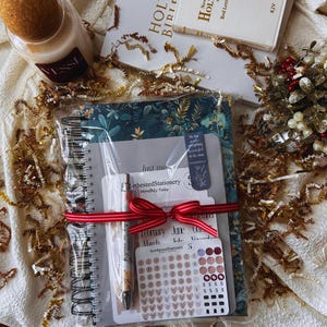 Puede incluir: Un juego de regalo con un diario floral, bolígrafo, pegatinas y una vela. El diario está encuadernado en espiral y atado con una cinta roja. El conjunto incluye una Biblia y elementos decorativos, ideal para regalos.