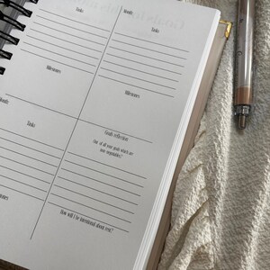 May include: Open planner with lined pages for tasks, milestones, and goal reflection. The planner is open to a spread with prompts and a pen rests on the side. The planner is beige with a spiral binding.