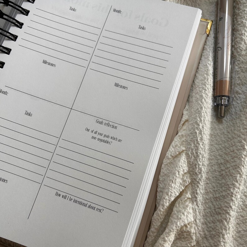 May include: Open planner with lined pages for tasks, milestones, and goal reflection. The planner is open to a spread with prompts and a pen rests on the side. The planner is beige with a spiral binding.