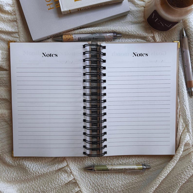 May include: An open notebook with lined pages and a black spiral binding. The words "Notes" are printed at the top of the pages. Several pens and a candle are arranged around the notebook, suggesting a workspace or study area.