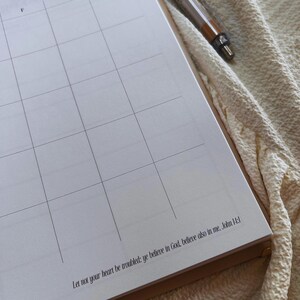 May include: A partially visible open notebook with a grid pattern, likely for scheduling or note-taking. The text "Let not your heart be troubled: ye believe in God, believe also in me. John 14:1" is printed at the bottom. A pen rests on the side.