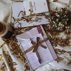 May include: A collection of religious and stationery items, including a Holy Bible, notebooks, and a pen. The Bible is white with gold lettering and a red letter edition. The notebooks are pink and white, and the pen is silver with a brown ribbon.