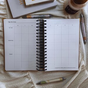 May include: An open, spiral-bound planner with a monthly calendar layout. The planner is gold-edged and lies on a cream-colored surface, with pens and a candle nearby. The calendar pages are blank, ready for use.