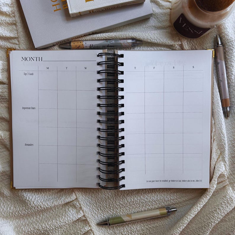 May include: An open, spiral-bound planner with a monthly calendar layout. The planner is gold-edged and lies on a cream-colored surface, with pens and a candle nearby. The calendar pages are blank, ready for use.