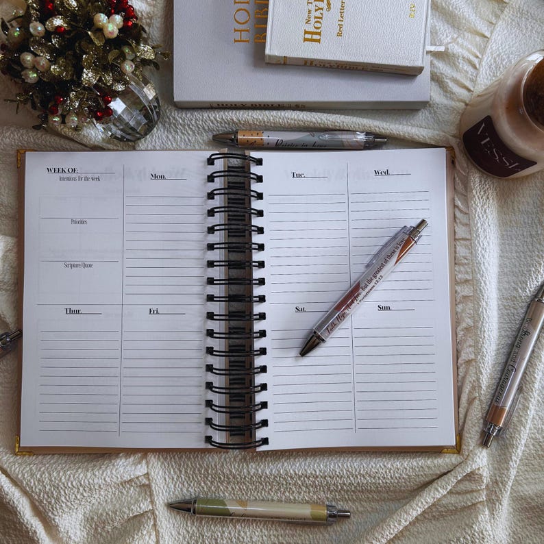 May include: Open weekly planner with lined pages, spiral binding, and gold trim. Several pens are scattered around the planner. The planner is open to a blank page with sections for each day of the week. Other items include books and a candle.