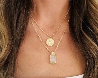 May include: A gold layered necklace with two pendants. The top pendant is a round coin with an intricate design. The bottom pendant is a rectangular piece of clear quartz.