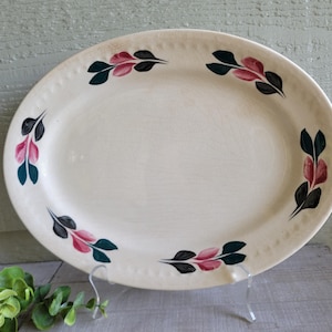 May include: An oval, off-white serving platter with a decorative border of small dots. The platter is adorned with a repeating pattern of stylized leaves in black, pink, and teal. The vintage-style dish is displayed on a clear stand.