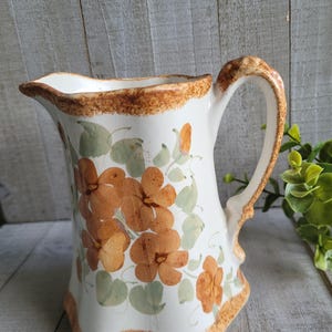 May include: A ceramic pitcher featuring a white base with hand-painted orange flowers and light green leaves. The rim and base are accented with a textured, mottled brown sponge-painted border. The pitcher has a curved handle and a pouring spout.