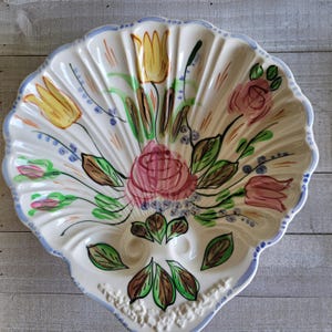 May include: A white, shell-shaped ceramic serving dish with a scalloped edge and a blue border. The dish is decorated with a floral design featuring pink roses, yellow tulips, and green leaves. The dish has two egg-shaped indentations.