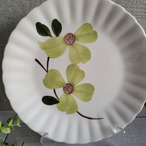 May include: White ceramic plate with a ruffled edge, featuring a hand-painted floral design. The flowers are light green with brown centers and dark green leaves. The plate is displayed on a clear stand, showcasing its decorative floral pattern.