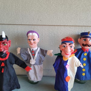 May include: Four vintage hand puppets. A red devil with horns and a black robe, a woman in a gray dress, a jester in blue and white, and a policeman in a blue uniform. The puppets are on glass bottles.