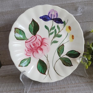 May include: Cream-colored, scalloped-edge plate with a hand-painted floral design. The design features a large pink rose, purple and blue irises, and yellow flowers with green leaves. The plate is displayed on a clear acrylic stand.