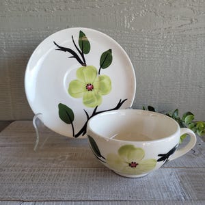 May include: White teacup and saucer set with a floral design. The cup and saucer feature a light green flower with a red center, black stems, and green leaves. The set is on a light gray wooden surface.