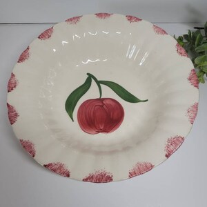 May include: A cream-colored ceramic serving bowl with a scalloped edge. The bowl features a hand-painted red apple design with green leaves in the center. The rim is decorated with red brushstrokes. A decorative piece for the kitchen.