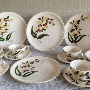 May include: A vintage white ceramic dinnerware set featuring a floral design in shades of green, brown, and yellow. The set includes plates, bowls, and teacups with saucers. The plates have a diameter of approximately 10 inches.