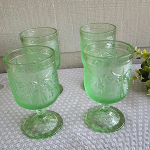May include: Four green-tinted glass goblets with a decorative floral pattern. The goblets have a rounded bowl shape, a short stem, and a circular base. The glass has a textured appearance, and the goblets are arranged on a white patterned surface.
