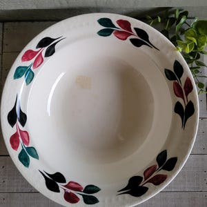 May include: Off-white ceramic bowl with a leaf pattern in black, red, and green around the rim. The bowl has a wide, shallow design, suitable for serving. The background is a light-colored wooden surface.