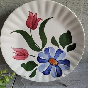 May include: White ceramic plate with a ruffled edge, hand-painted with a floral design. The design features a red tulip, a blue daisy, and green leaves. The plate is displayed on a clear stand.
