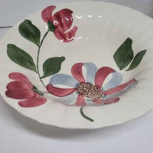 May include: A white ceramic serving bowl with a scalloped edge, adorned with hand-painted floral designs. The flowers feature burgundy and light blue petals, green leaves, and a textured center.