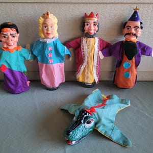 May include: A collection of vintage hand puppets, including a witch, princess, king, magician, and a dragon. The puppets feature painted faces and fabric clothing in various colors like teal, purple, pink, and orange. The dragon puppet is lying flat.
