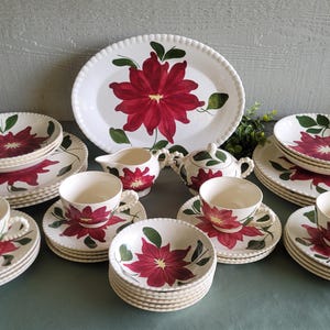 May include: A complete vintage porcelain dinnerware set featuring a red poinsettia floral design on a white background. The set includes plates, bowls, cups, saucers, a serving platter, a creamer, and a sugar bowl.