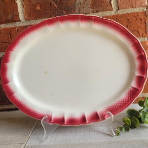 May include: An oval serving platter with a white center and a red, scalloped border. The border has a textured, diamond-like pattern. The platter is displayed on a clear stand against a brick wall.