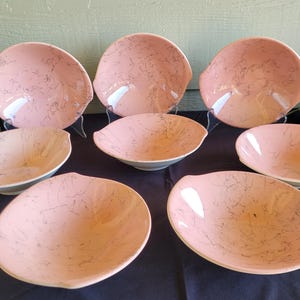 May include: A set of eight vintage pink ceramic bowls with a speckled design. The bowls have a unique shape with a slightly raised rim and are arranged on a black surface.