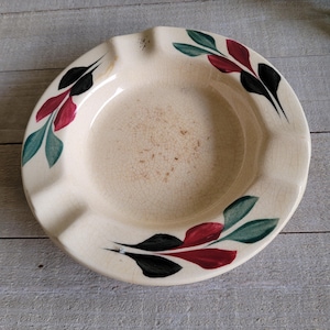 May include: Cream-colored ceramic ashtray with a leaf design in black, red, and teal. The ashtray has a circular shape with four notches for resting cigarettes. The interior shows signs of use.