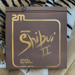 May include: A brown portfolio with gold lettering and a handle. The text on the front reads "2M presents Shibui II", with the designers' names below. The portfolio is square and has a leather handle.