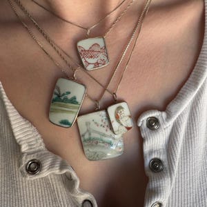 May include: Four delicate necklaces with rectangular and square pendants. Each pendant features a unique hand-painted design in red, green, and blue on a white background. The pendants are framed in silver and hang on thin silver chains.
