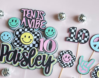 May include: A set of 10 cake toppers featuring a black and white checkered pattern with smiley faces, peace signs, and the text "Ten is a vibe Paisley 10".