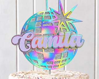 May include: A cake topper featuring a silver glitter disco ball with an iridescent finish and a starburst accent. The name Camila is written in a purple script font across the center. The topper is displayed on a white frosted cake.