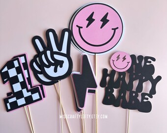 May include: Set of six black and pink paper cake toppers with various designs, including a peace sign, a lightning bolt, a checkered number one, and two smiley faces with lightning bolts for eyes. The text "One Happy Babe" is also included.