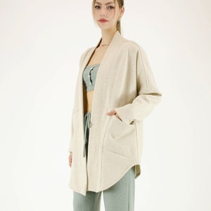 May include: A long-sleeved, beige cardigan with a relaxed fit and a curved hem. The cardigan has a shawl collar and two pockets. The model is wearing the cardigan with a light green tank top and matching pants.