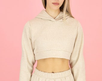 May include: A beige cropped hoodie and matching sweatpants. The hoodie has a drawstring hood and the sweatpants have a drawstring waistband and cargo pockets.