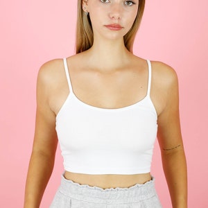 May include: A woman wearing a white crop top and gray sweatpants. She is standing in front of a pink background.