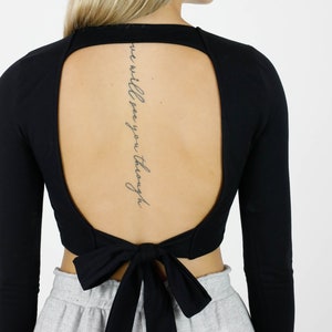 May include: Black long-sleeve top with an open back design and a bow tie closure. The top features a scoop neckline and long sleeves. The backless design reveals a tattoo with the words "we will see you through".