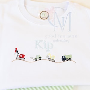 May include: A white t-shirt featuring an embroidered design of four construction vehicles including a red crane, a green tractor, a yellow excavator, and a green dump truck on a brown stitched line. The name Kip is embroidered in light blue.