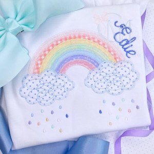 May include: A white onesie with a rainbow, clouds, and raindrops embroidered on it. The name "Edie" is embroidered in blue on the right side of the onesie.