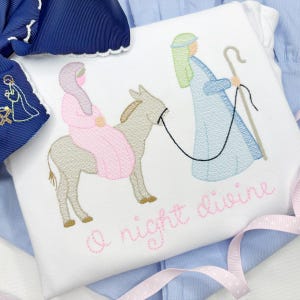 May include: White baby onesie with an embroidered nativity scene. The design features a woman riding a donkey, a figure in blue, and the words "A night divine" in pink script. A blue bow and ribbon are in the background.