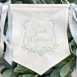 May include: A white banner with blue embroidery that says "Just Married" in a decorative script font. The banner has a blue ribbon on each side and is decorated with a white bow and green vines.