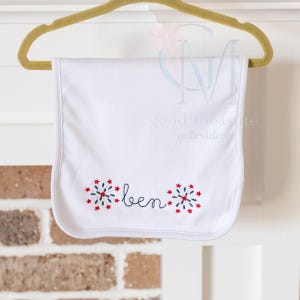 May include: White baby bib with the name "ben" embroidered in a cursive font, flanked by red and blue starburst designs. The bib is hanging on a gold hanger.