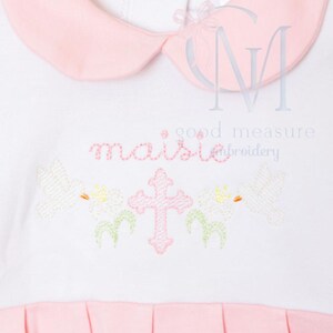 May include: White dress with a pink collar and hem. The dress features embroidered details including a pink cross, flowers, and the word "maisie" in cursive. The words "good measure embroidery" are also visible.