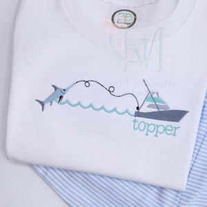 May include: White long-sleeved shirt with an embroidered fishing scene. A light blue marlin is hooked to a fishing line connected to a gray boat with the word "topper" in light blue. Blue and white striped pants are visible.